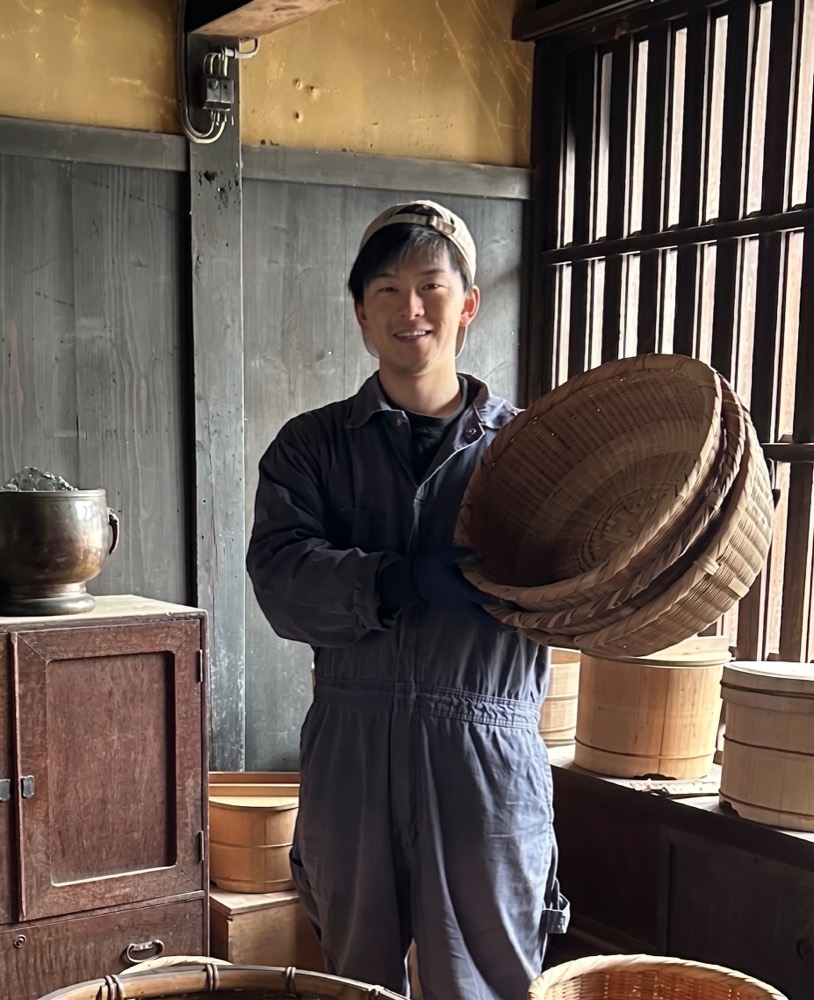 代表社員 永田大樹。土蔵の中で竹籠を手にした作業着姿のポートレート