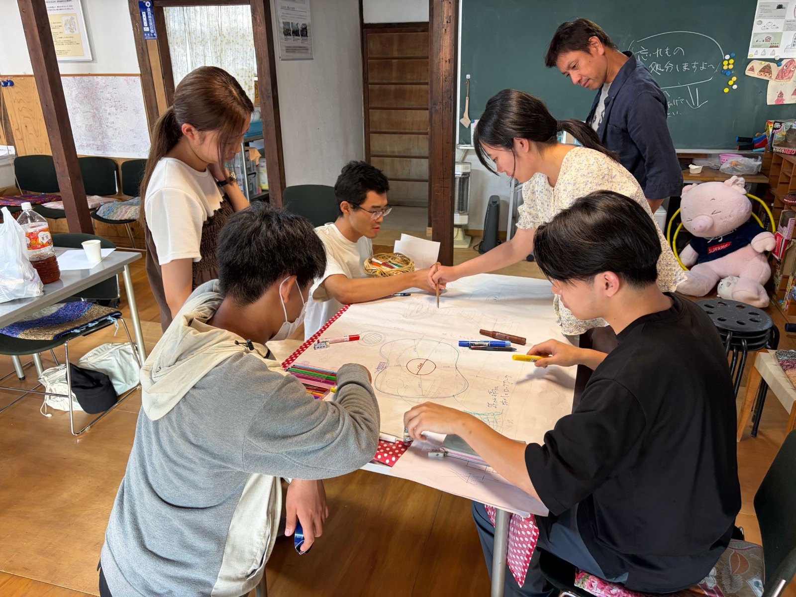 古い建物の一室で、大きな紙を囲んでツリーハウスの構想を描き込む学生たち